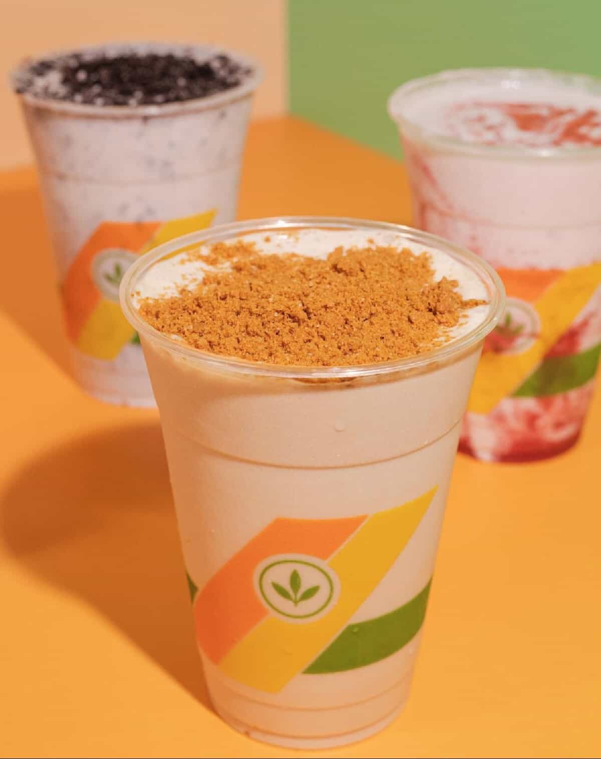 Three Plant Power Fast Food milkshakes in clear plastic cups with different toppings on an orange countertop with a green background.