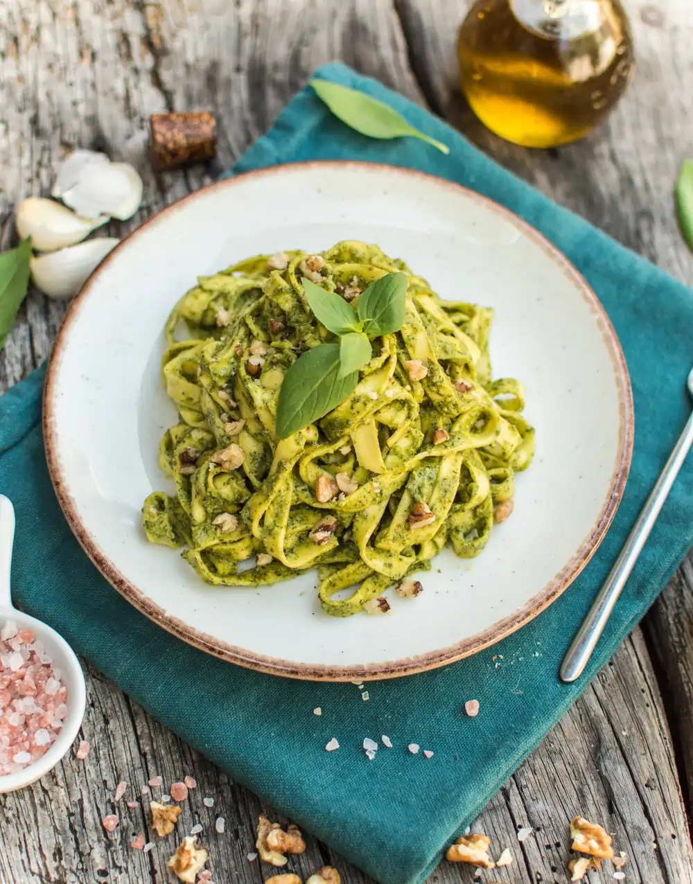A neutral dish of vegan walnut pesto pasta with some decorative basil and some cloves of garlic on the side 
