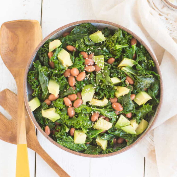 A bowl of cooked kale salad with beans, diced avocado, and sesame seeds.