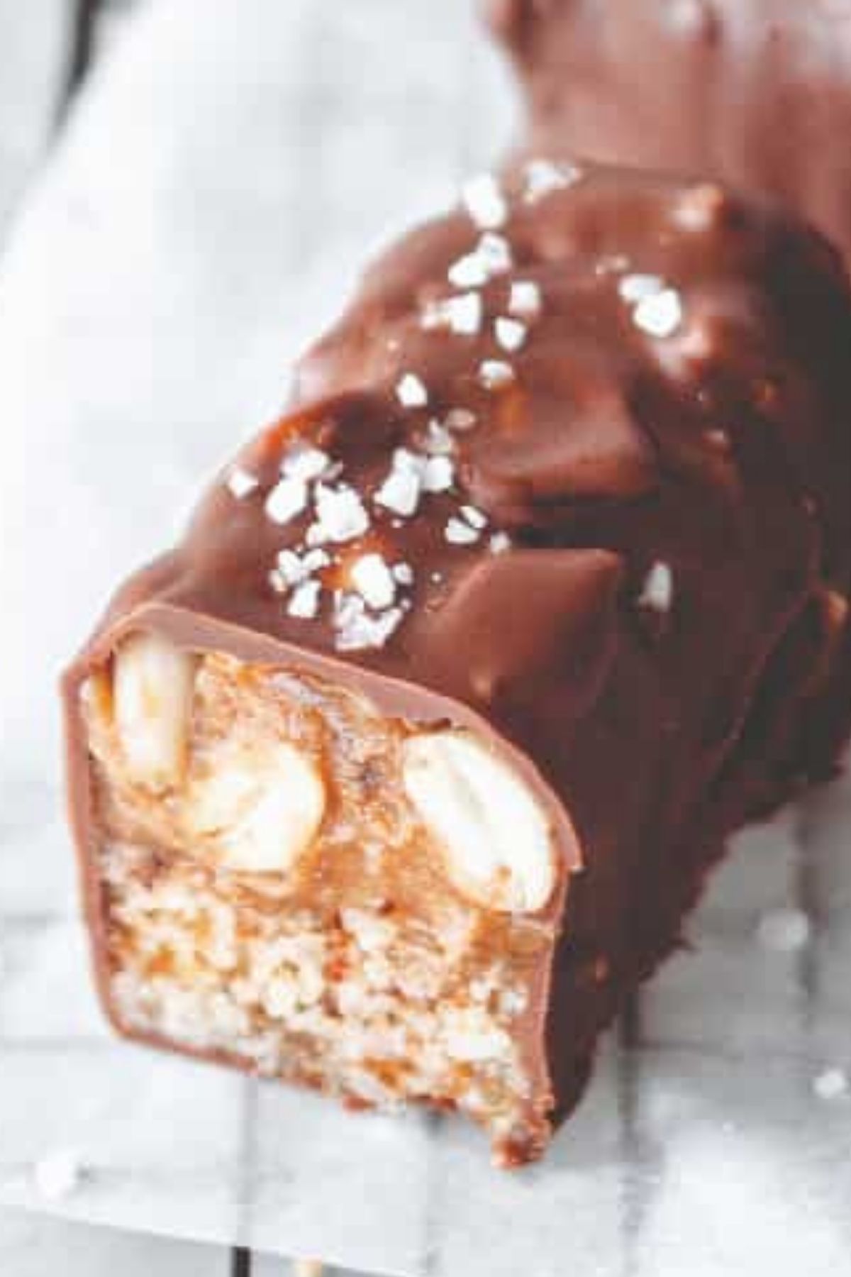 Close-up of a homemade vegan Snickers bar with a thick chocolate shell, gooey caramel filling, and crunchy peanut base, topped with flaky sea salt.