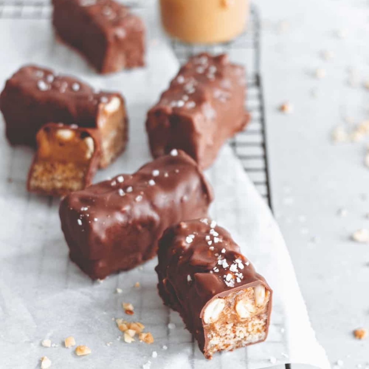 Homemade vegan Snickers bars on parchment paper, coated in rich chocolate and sprinkled with flaky sea salt. A bite reveals chewy caramel and crunchy roasted peanuts inside.