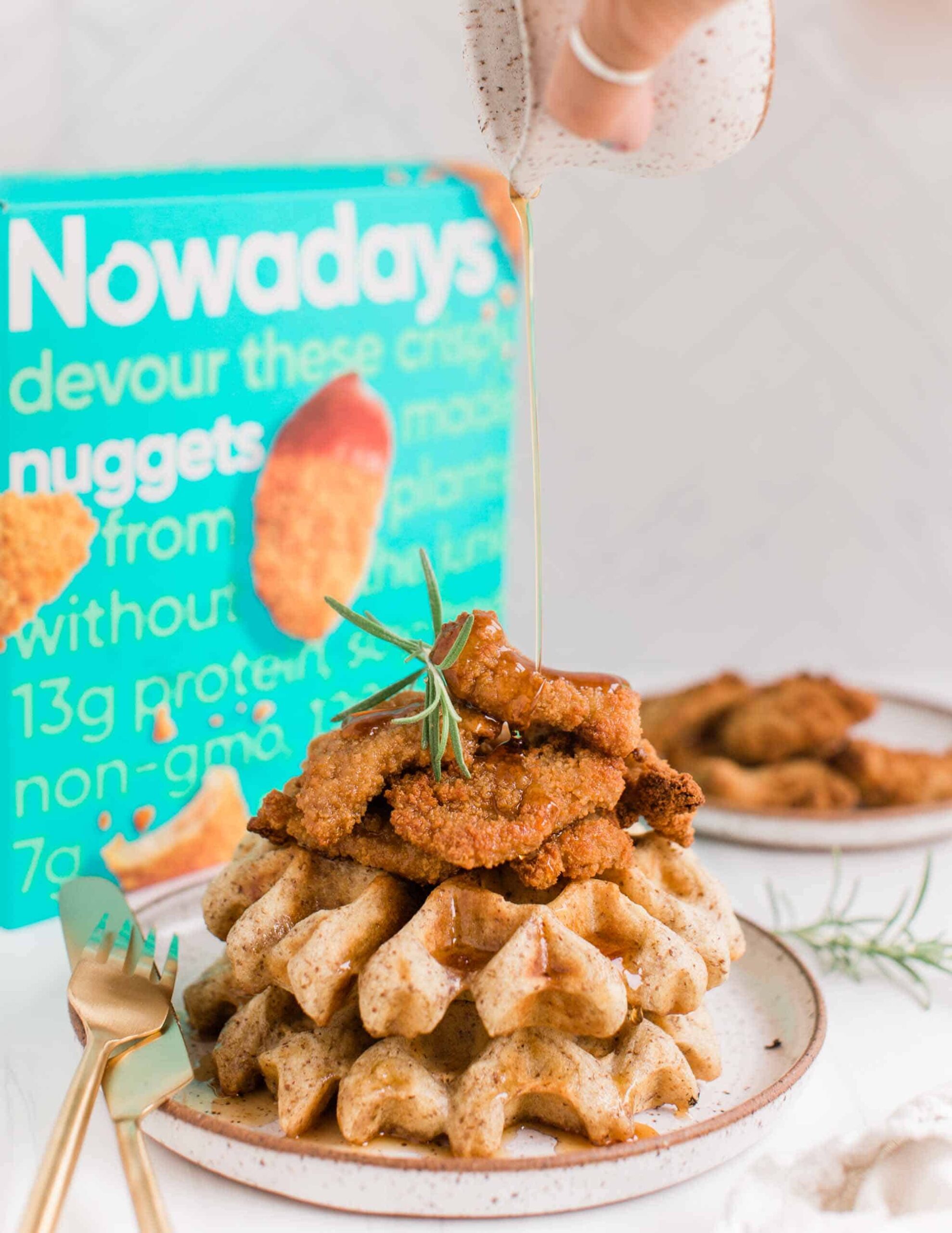 Vegan Chicken and Waffles Stacked on a Plate With a Hand Pouring Maple Syrup on Top in front of a Nowadays Vegan Nuggets Box