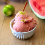Three scoops of watermelon gelato in a ramekin with a sliced watermelon and lime in the background.