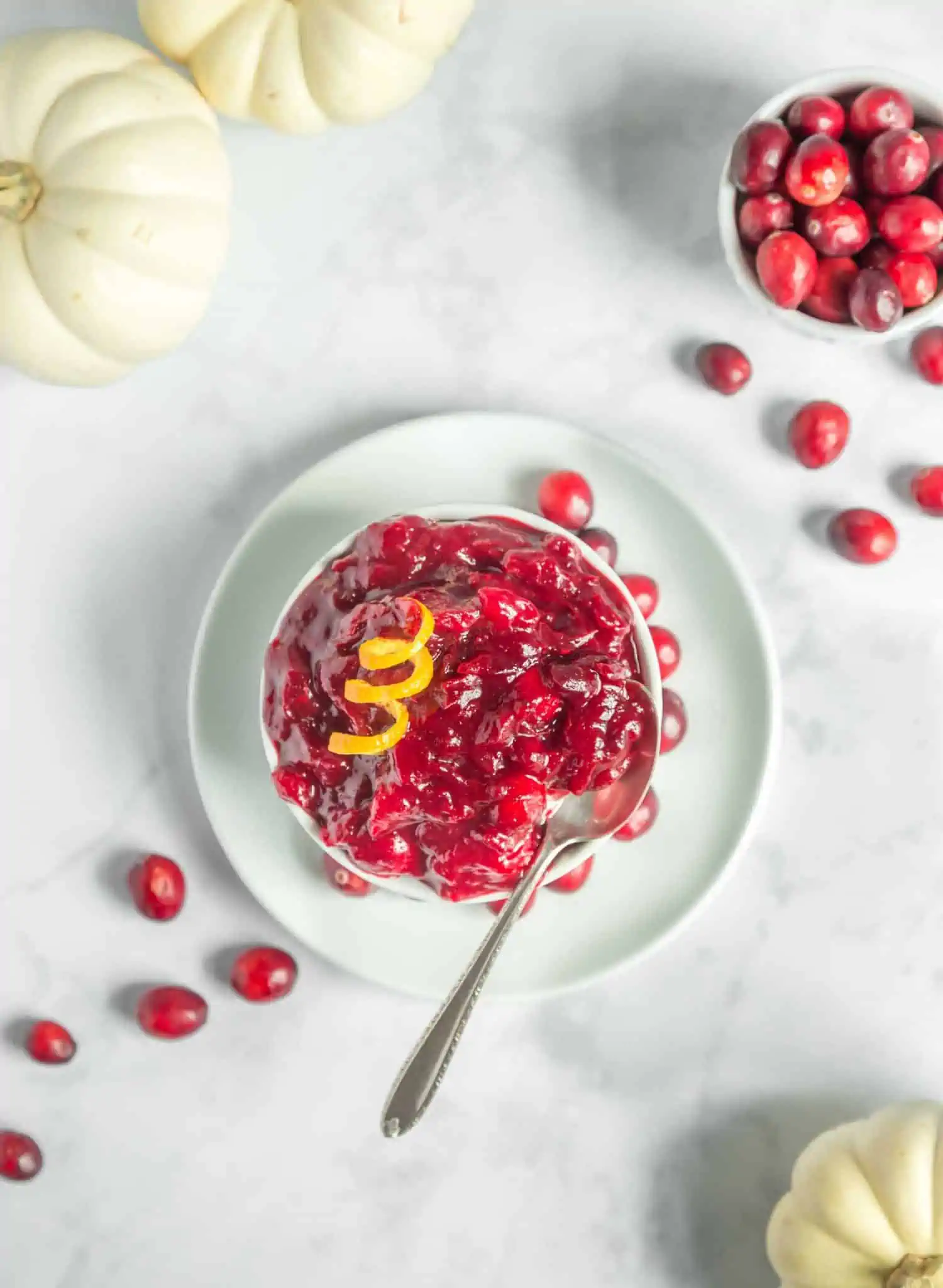 Easy vegan homemade cranberry sauce with orange juice and agave in a white bowl with spoon and scattered cranberries. 
