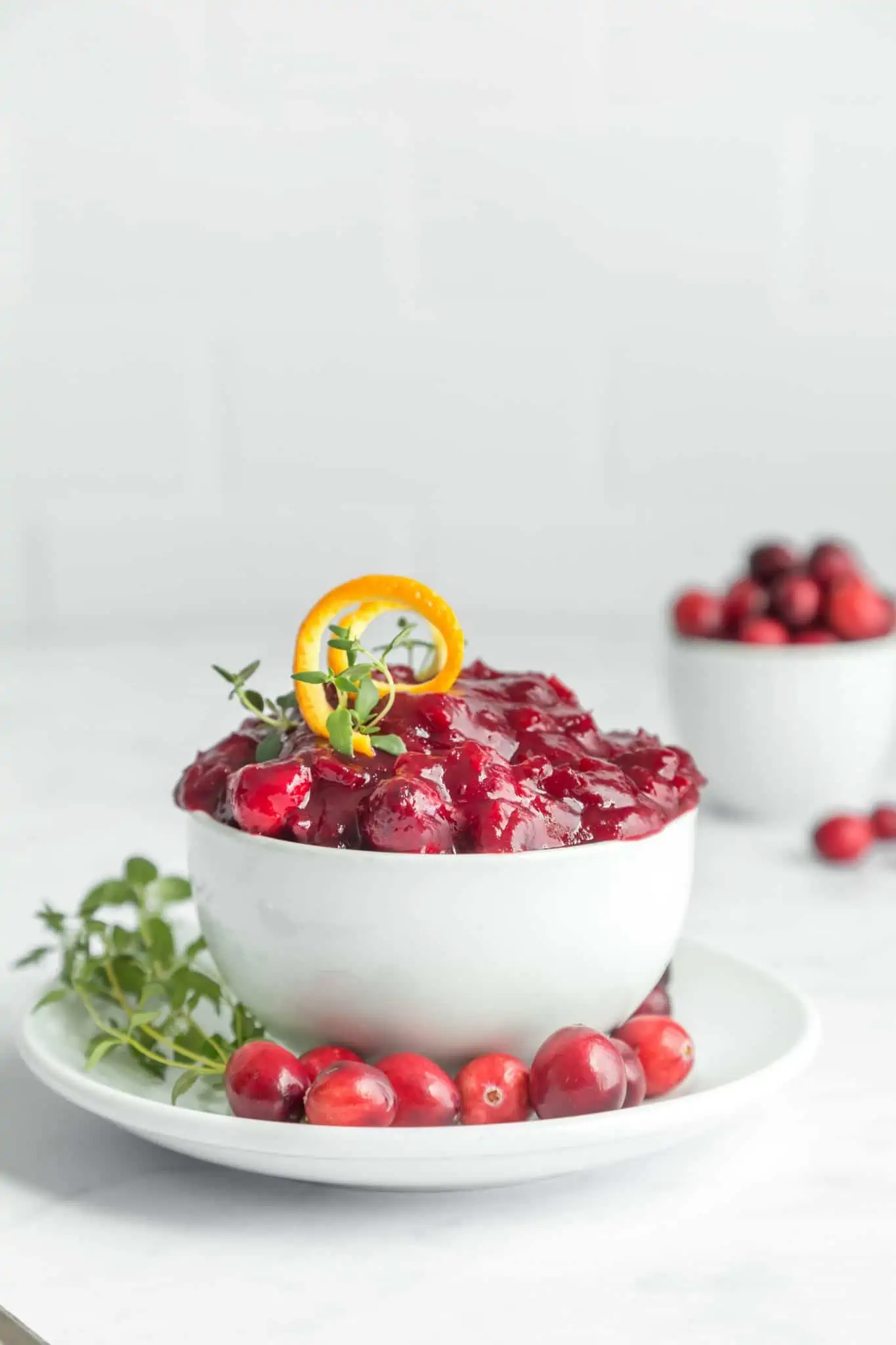 Vegan Thanksgiving recipe for homemade cranberry sauce. Pictured in a white bowl and saucer along with fresh greens, cranberries and orange zest. 
