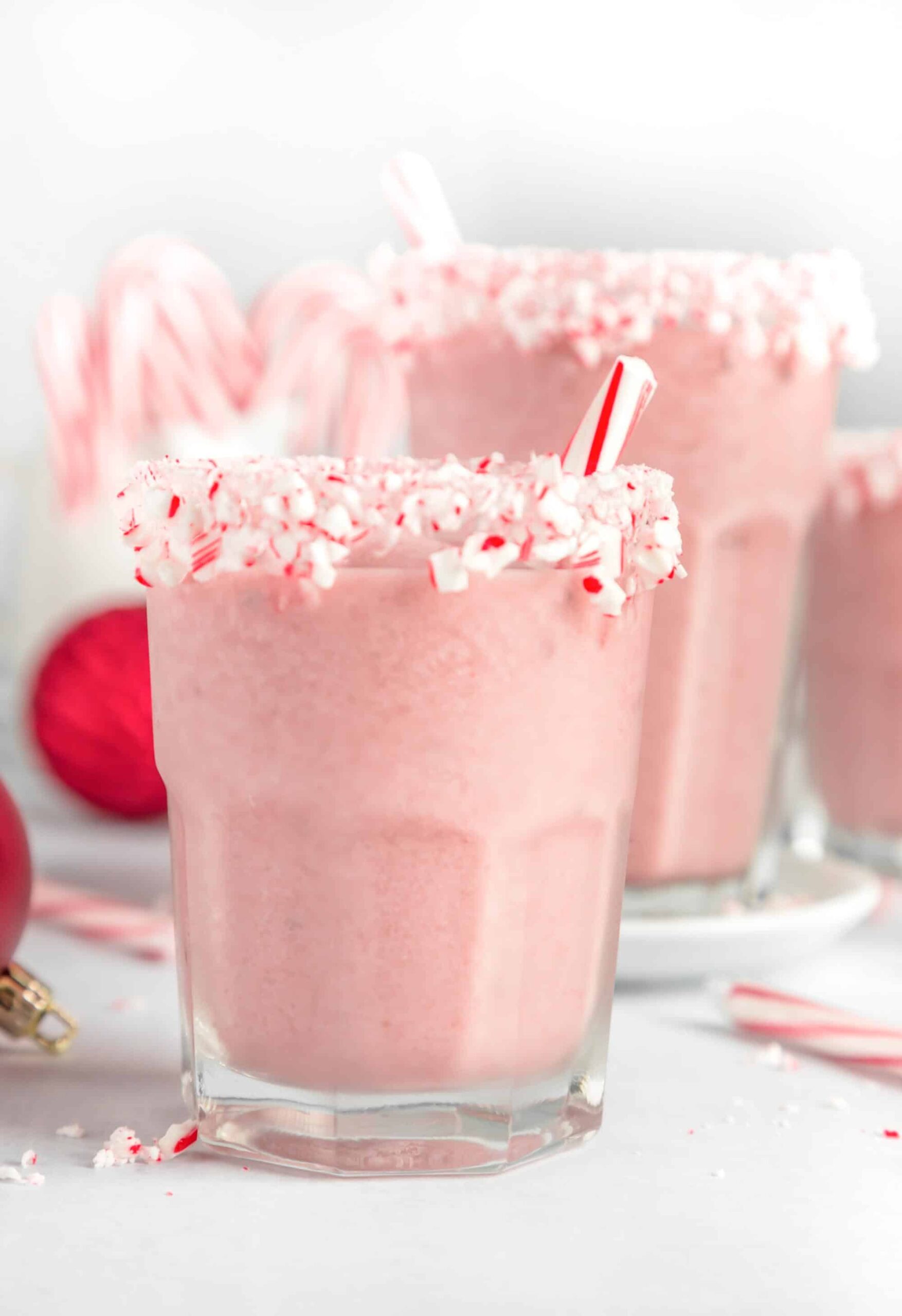 A healthy pink peppermint smoothie recipe for the holidays in a glass with crushed candy cane on the rim and a hollow peppermint stick for stirring. 