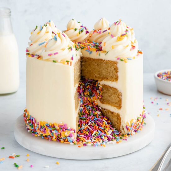 Classic three layer vegan cake decorated with vanilla frosting and rainbow sprinkles spilling out of it.