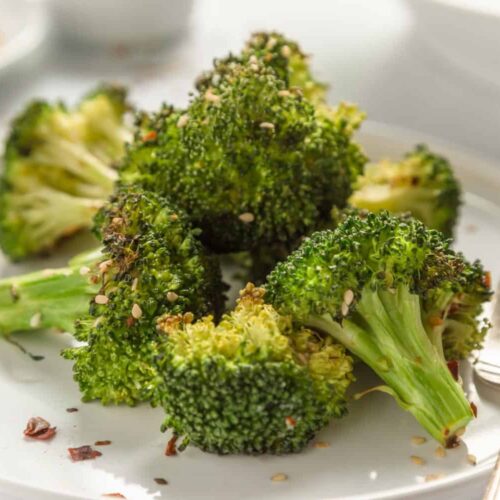 Air fried broccoli sprinkled with sesame seeds served on a white plate with a rustic fork