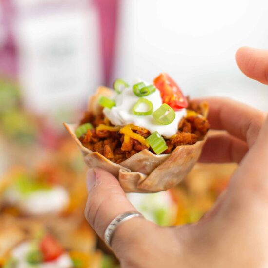 A hand holding up a vegan taco cup topped with vegan sour cream and sliced green onions.