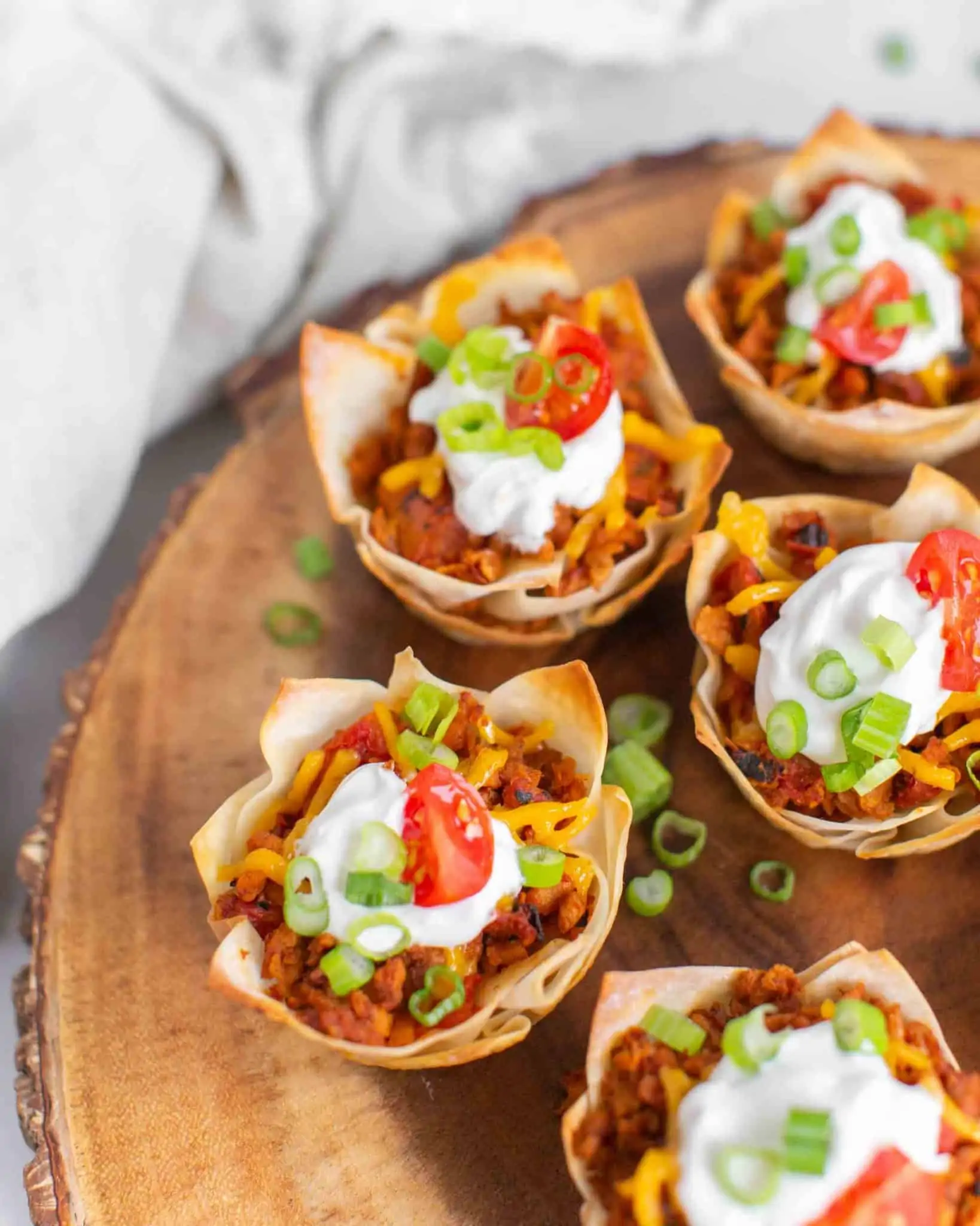 Vegan wonton taco cups served on a wooden platter and topped with vegan sour cream, sliced green onion and cherry tomatoes.