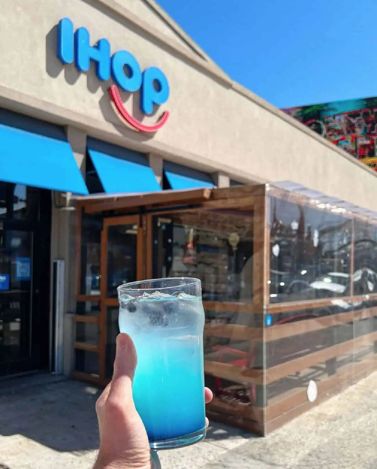 A hand holding a glass filled with blue colored Coney Island Lemonade in front of an IHOP restaurant location. 