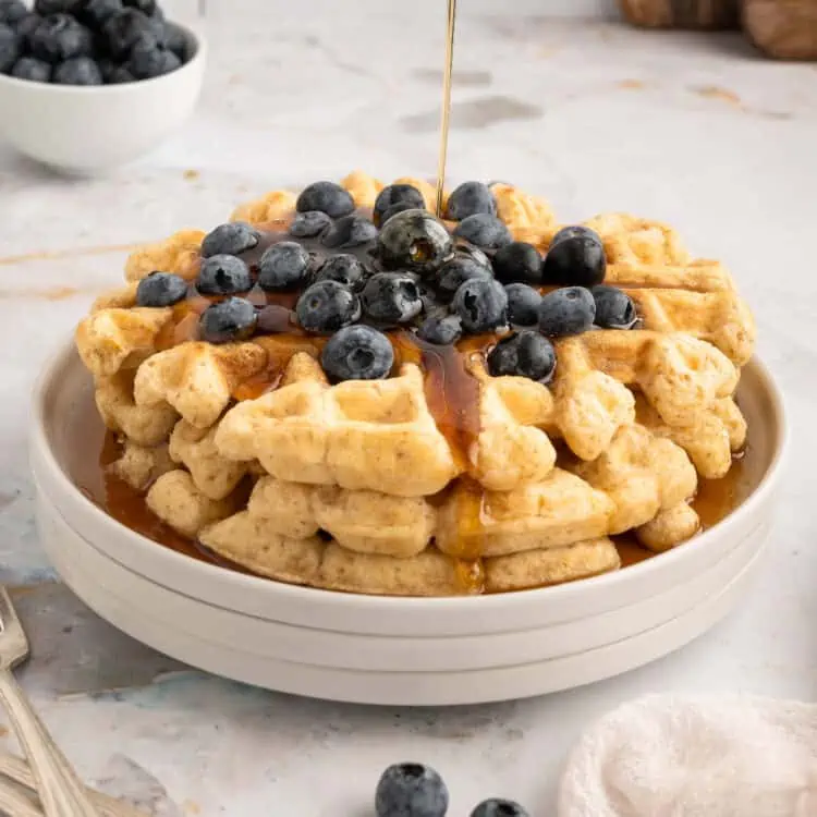 A stack of vegan waffles, topped with blueberries and drizzled with syrup.