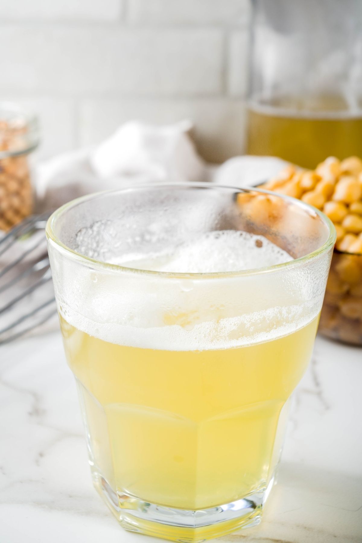 Glass filled with aquafaba, the yellowish liquid drained from canned chickpeas, set on a white countertop with chickpeas and a whisk in the background.