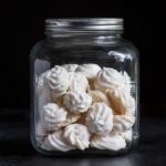 Crisp white vegan meringue cookies stacked in a clear glass jar with a silver lid, showing off their light and airy texture.