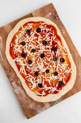 Vegan pizza dough rolled out flat topped with red sauce, dairy-free cheese, and black olives.