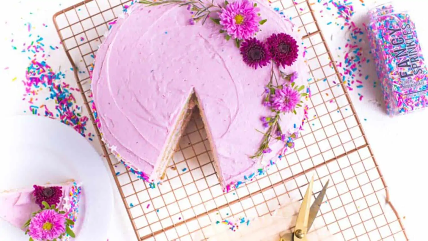 vegan birthday cake with pink frosting, sprinkles, and flowers on a gridded surface