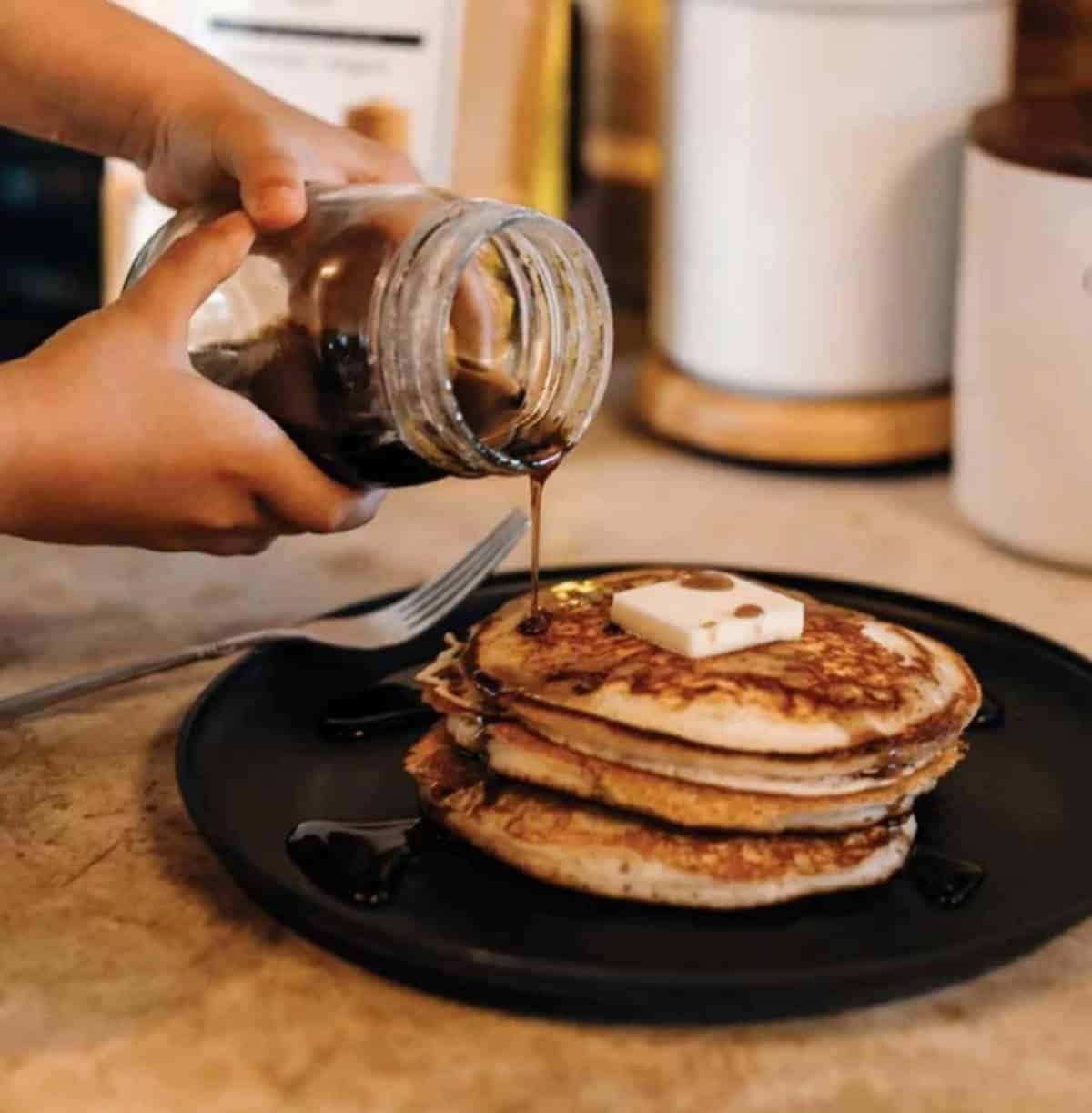 Hands holding a jar of syrup and pouring it over a stack of vegan pancakes on a plate.
