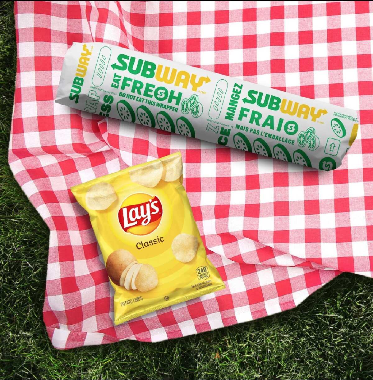 A wrapped Subway sandwich and a bag of Lay's potato chips laid out on red gingham blanket  on the grass. 