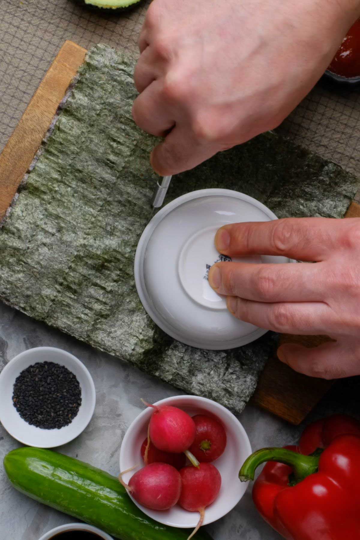 A soy sauce dish being used as a guide to cut pieces of nori into circles.