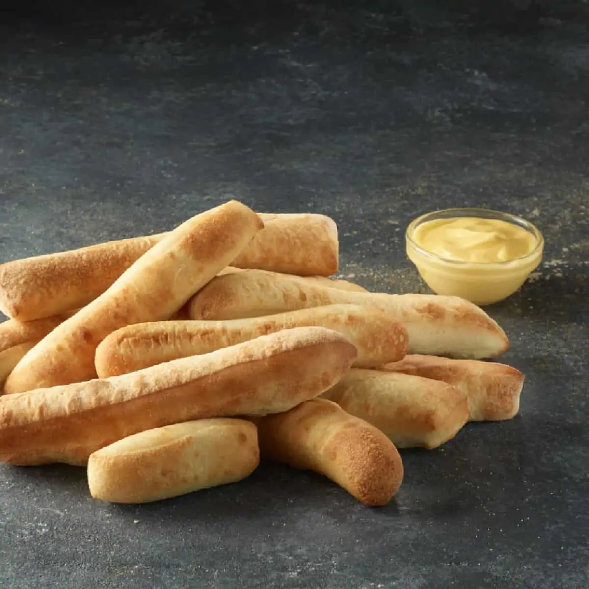 A pile of plain Papa John's breadsticks on a dark gray countertop with a small side of garlic sauce in a clear glass bowl to the right.