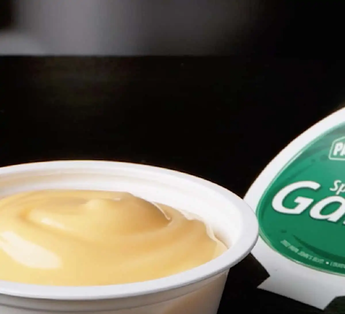 A small white container of pale yellow Papa John's garlic sauce with lid to the right on a black background.