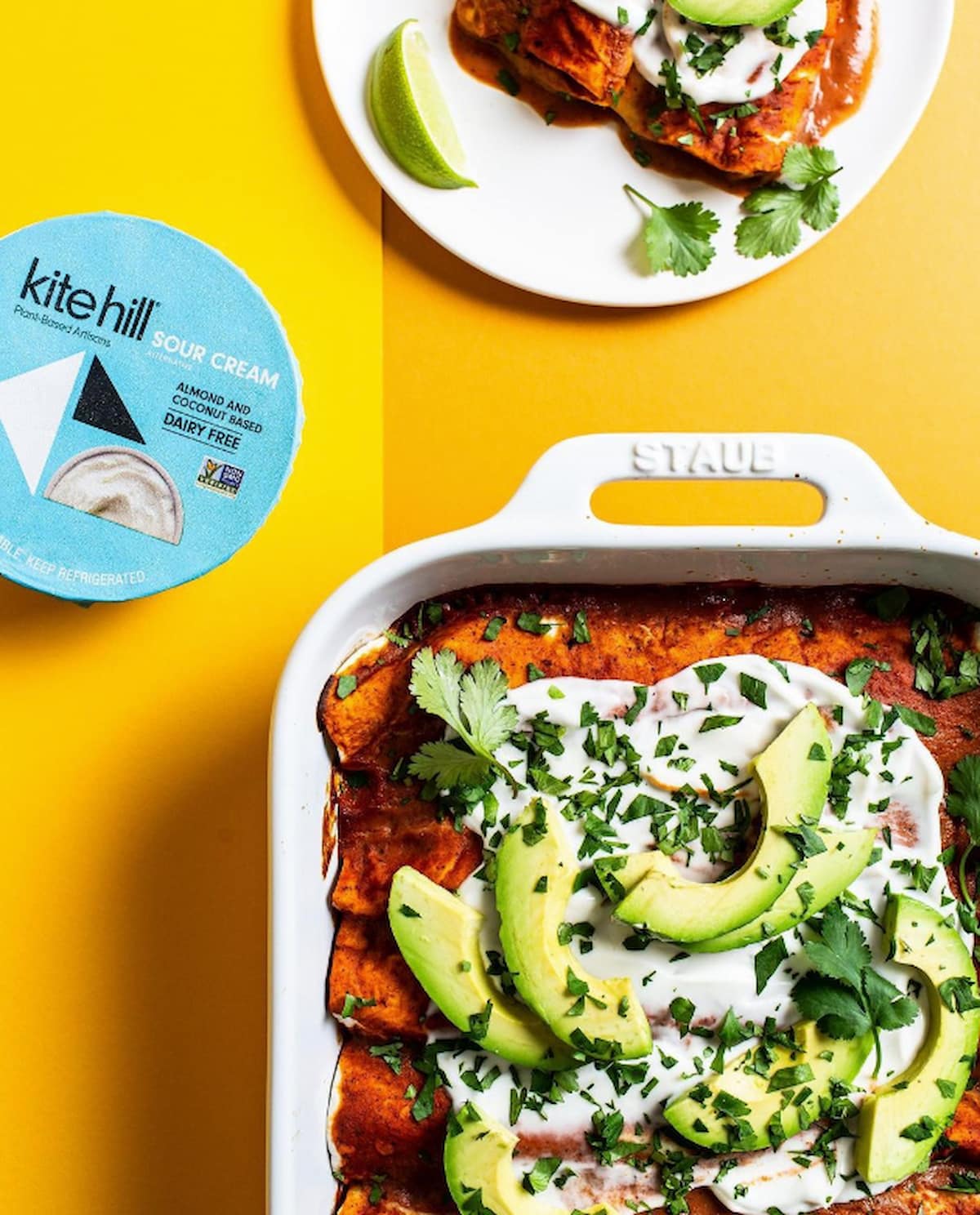 Kite Hill vegan sour cream container next to a casserole dish of plant-based enchiladas topped with vegan sour cream.