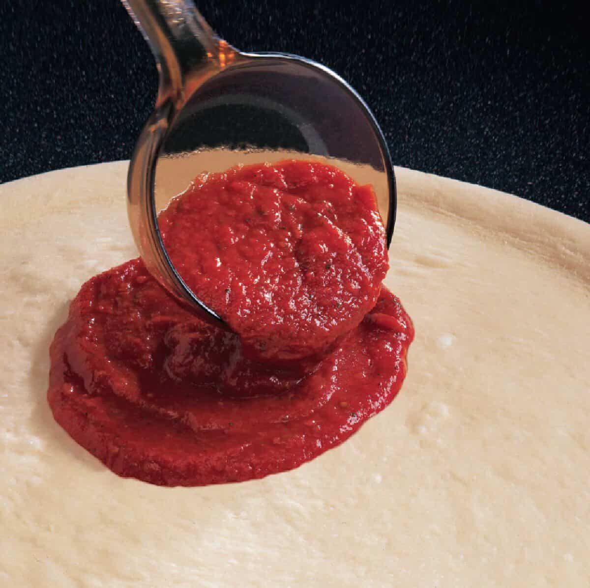 A scoop of Papa John's pizza sauce is being spread over an uncooked pizza dough circle.