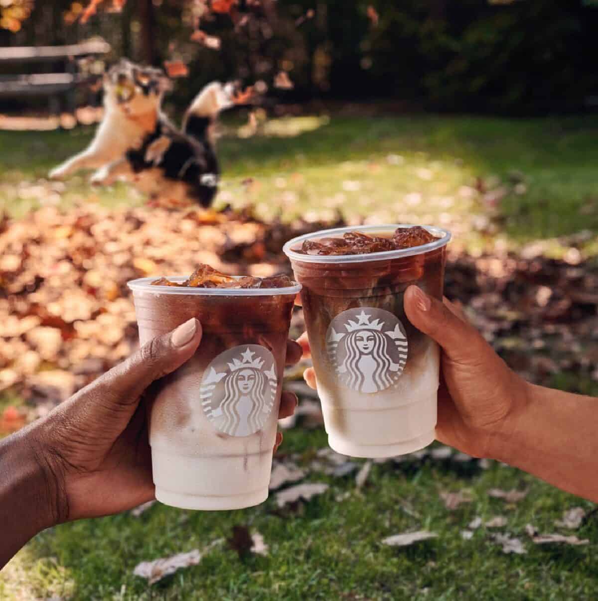 Two hands each holding a plastic cup of Starbucks Iced Apple Crisp Oatmilk Macchiatos outside on a fall day with leaves in the background.