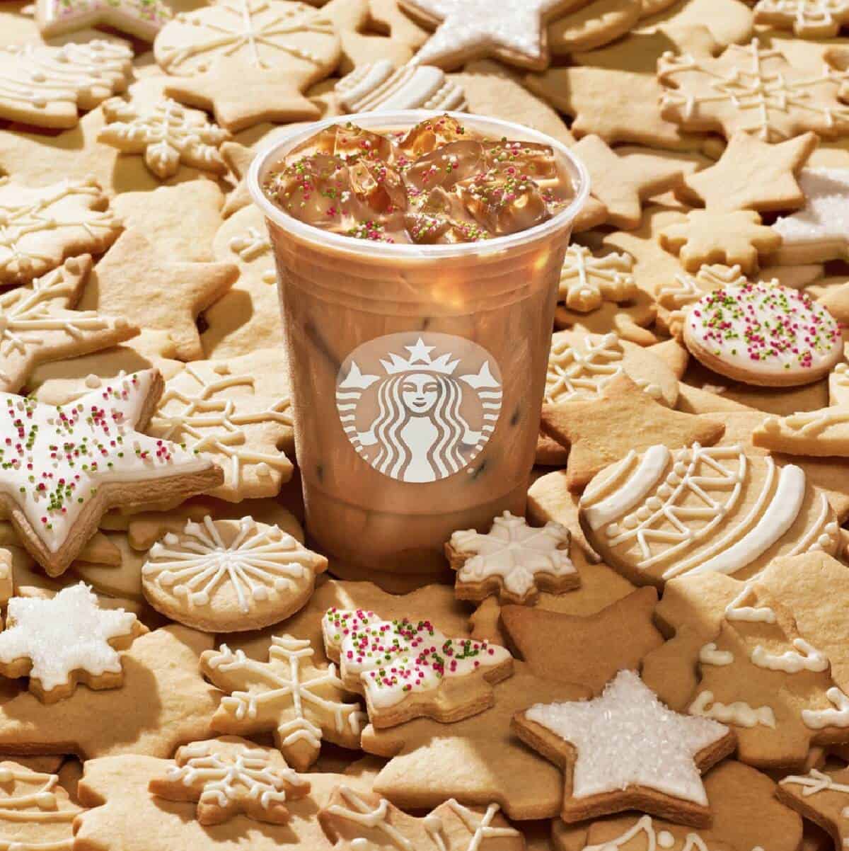 A clear plastic cup with Starbucks symbol filled with Iced Sugar Cookie Almondmilk Latte on top of a pile of white iced sugar cookies. 