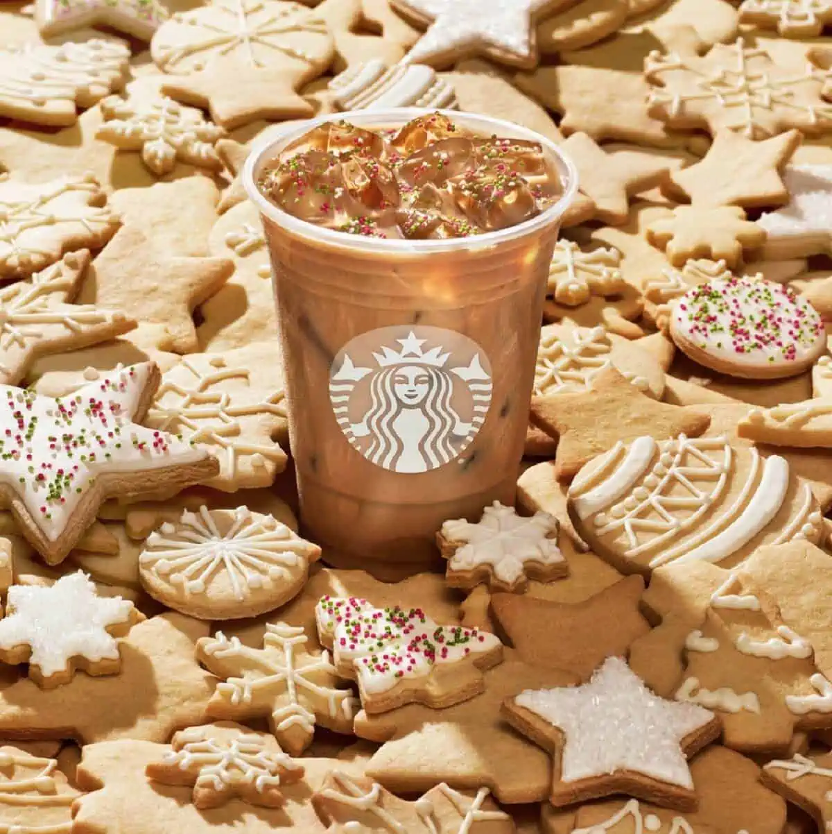A clear plastic cup with Starbucks symbol filled with Iced Sugar Cookie Almondmilk Latte on top of a pile of white iced sugar cookies. 