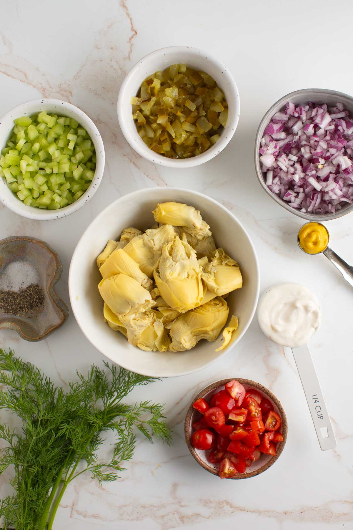 Artichoke tuna ingredients flatlay with artichokes, tomatoes, onion, pickles, celery, dill, vegan mayo, mustard, salt, and pepper. 