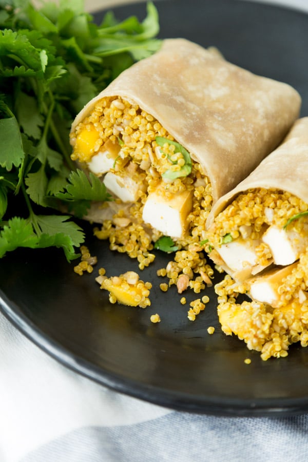 one wrap cut in half filled with curried quinoa and cooked tofu in a tortilla on a black plate with cilantro garnish