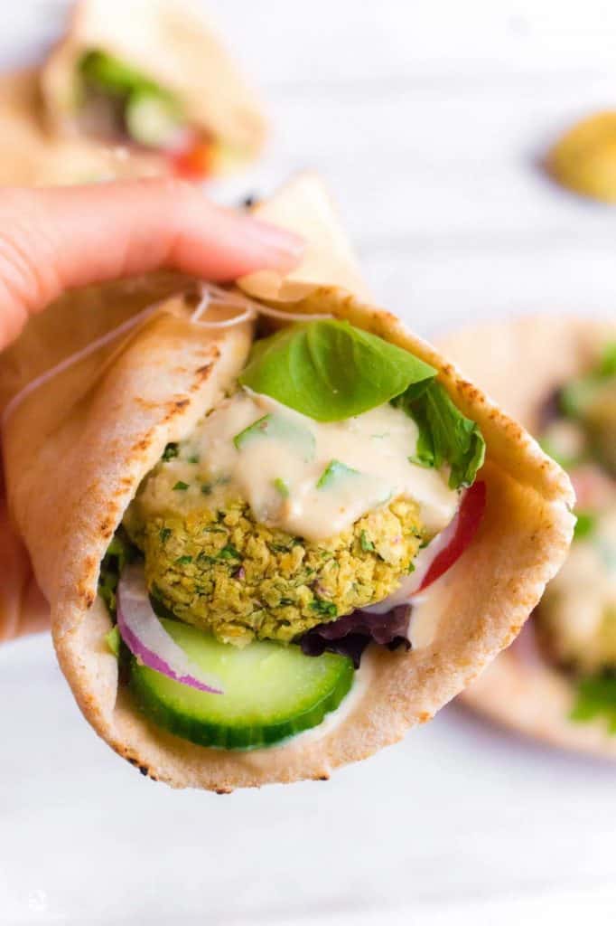 white hand holding a falafel wrappe in pita with vegan sauce, cucumber, red onion, and lettuce