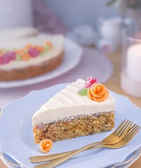 slice of carrot cake with vegan cream cheese frosting and marzipan roses on a white plate with two gold forks resting on the plate