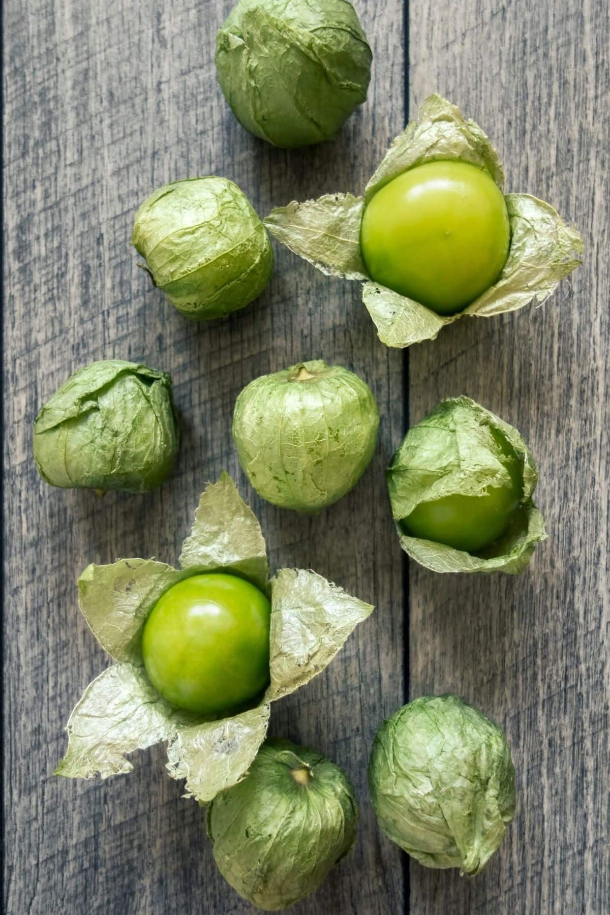 Fresh green tomatillos, some partially husked to reveal their shiny fruit, arranged on a rustic wooden surface.