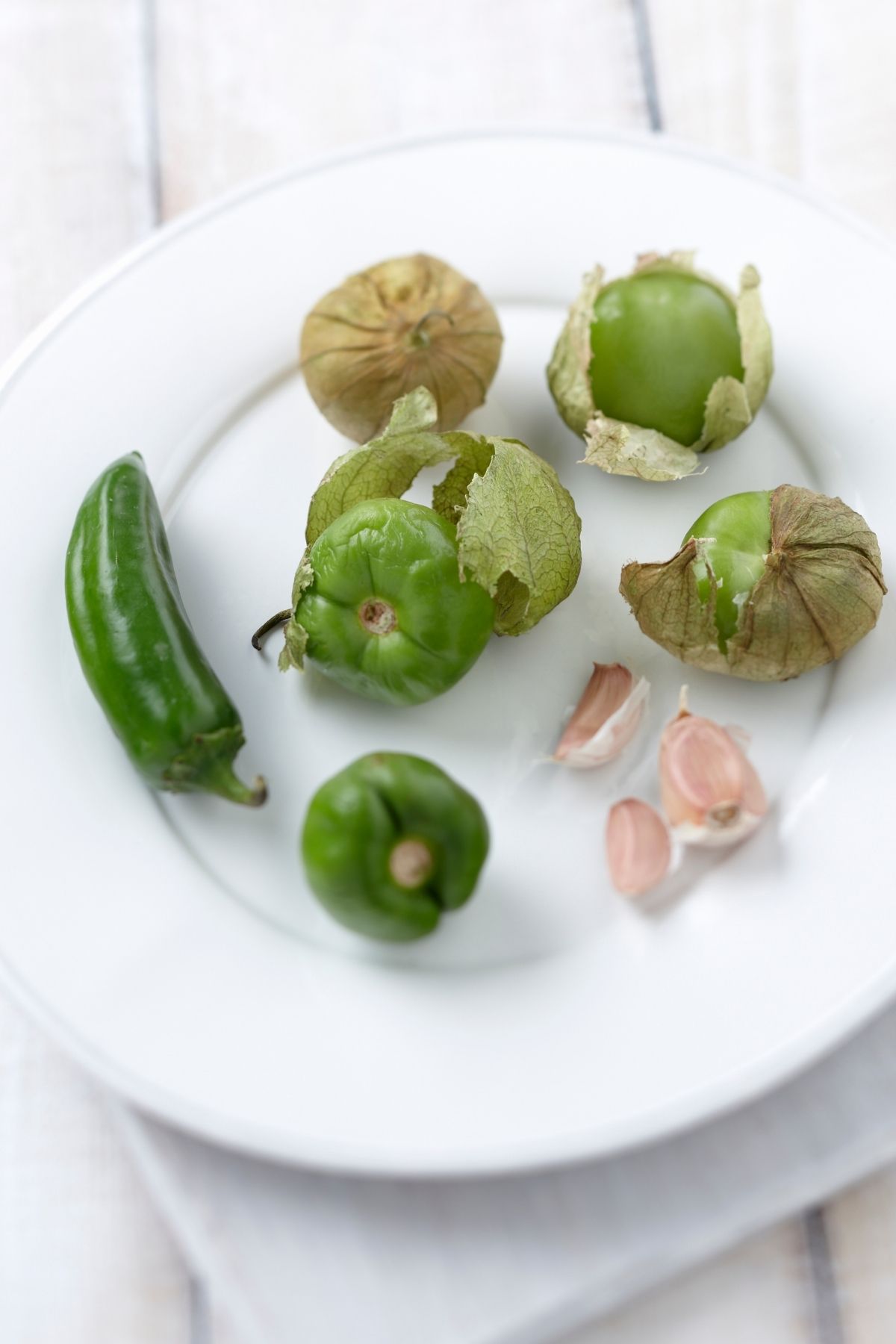 A white plate with tomatillos, jalapeno, and a few cloves of garlic.
