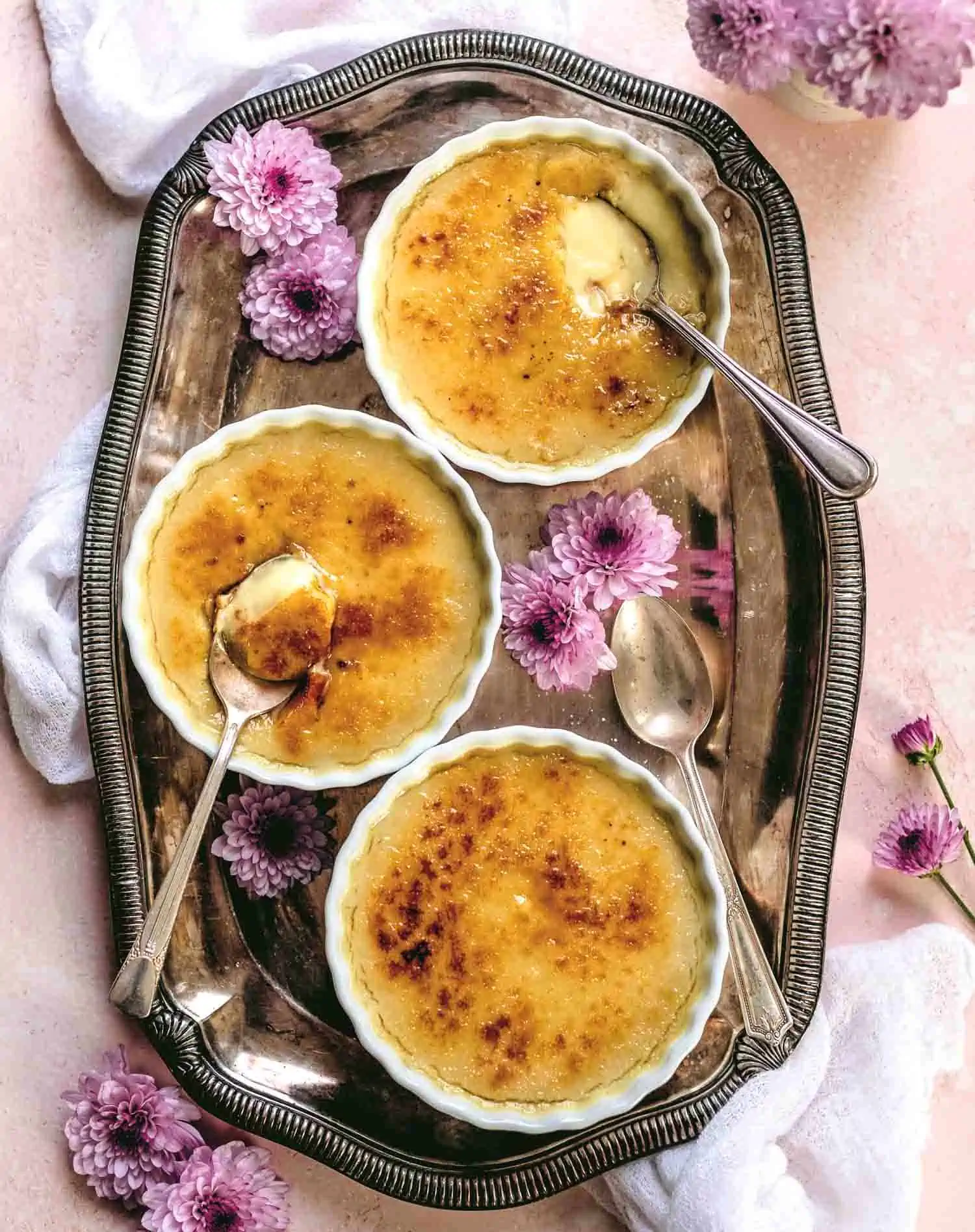 Serving platter with three vegan creme brulee ramekins with spoons scooped in them and delicate purple flowers around them. 