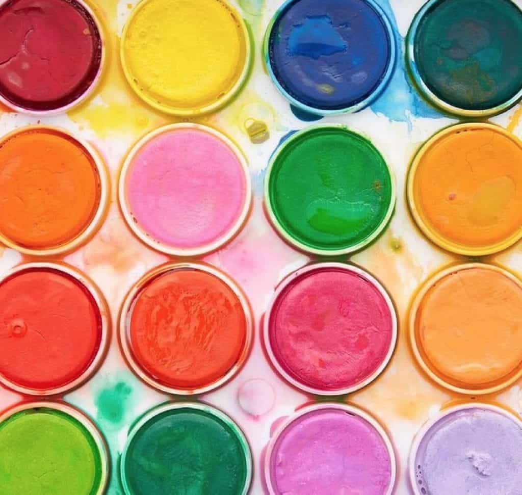 A four by four grid of rainbow colored circular paints in a white paint palette.