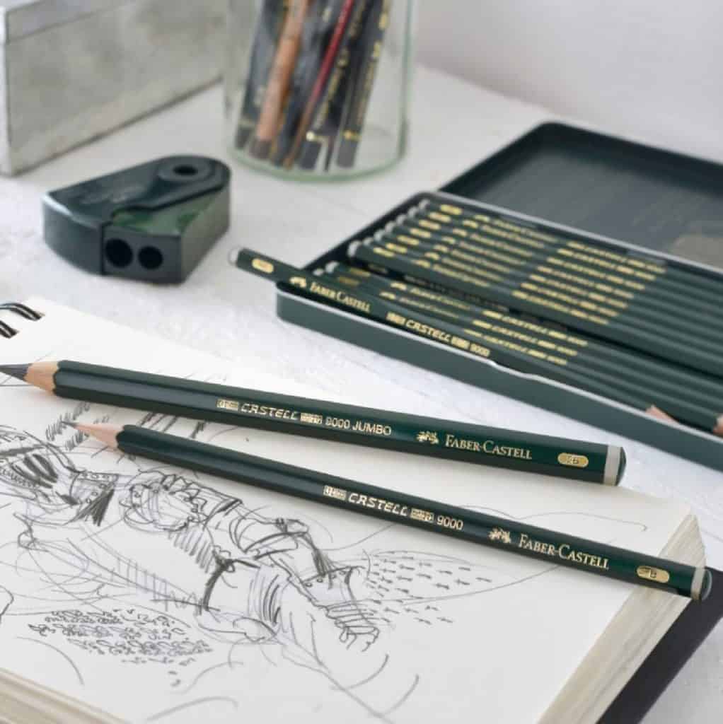 A sketchbook and pencil sharpener on a table with a set of dark green colored Faber-Castell cruelty-free pencils.