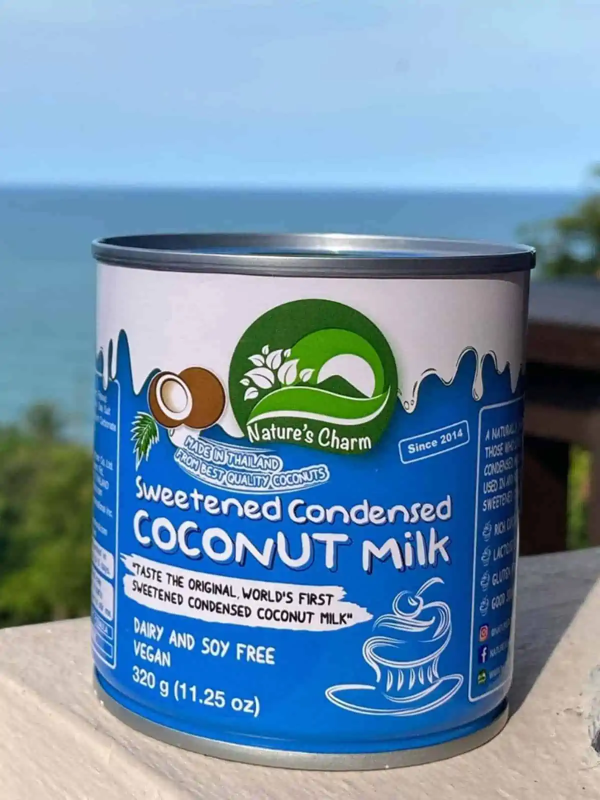 Can of Nature's Charm Sweetened Condensed Coconut Milk on sitting on wooden table with ocean and trees behind it.