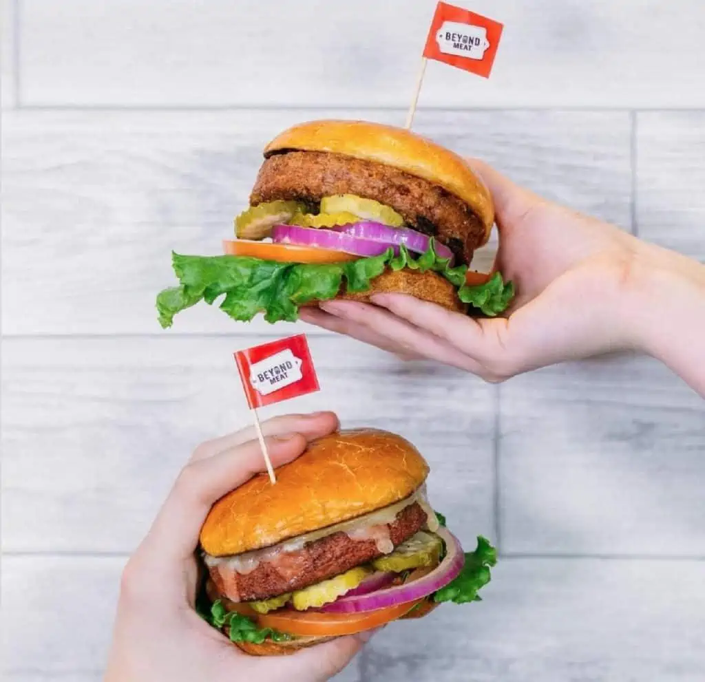 One hand on the right of the photo holding out a plant-based Beyond Meat burger with a little red flag and another hand doing the same thing coming up from the bottom of the photo. The background is a light gray wooden paneled wall.