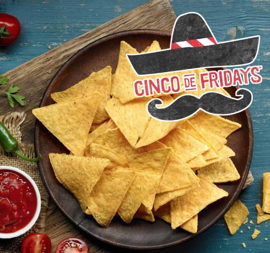 A wooden plate on top of a cutting board filled with corn tortilla chips and surrounded by a small dish of red salsa and green peppers and cilantro. It says "Cinco de Fridays" in red with a black hat and mustache.