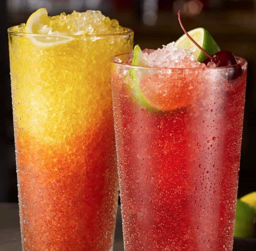 Two tall glasses filled with colorful fruit slushies. The slushie on the left is yellow and orange with a lemon wedge and the other red with lime wedges and a cherry.