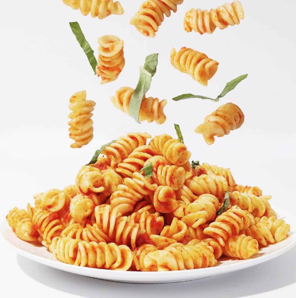 A white plate filled with vegan-friendly fusilli pasta and covered with tomato basil sauce. Pasta and basil leaping happily in the air.