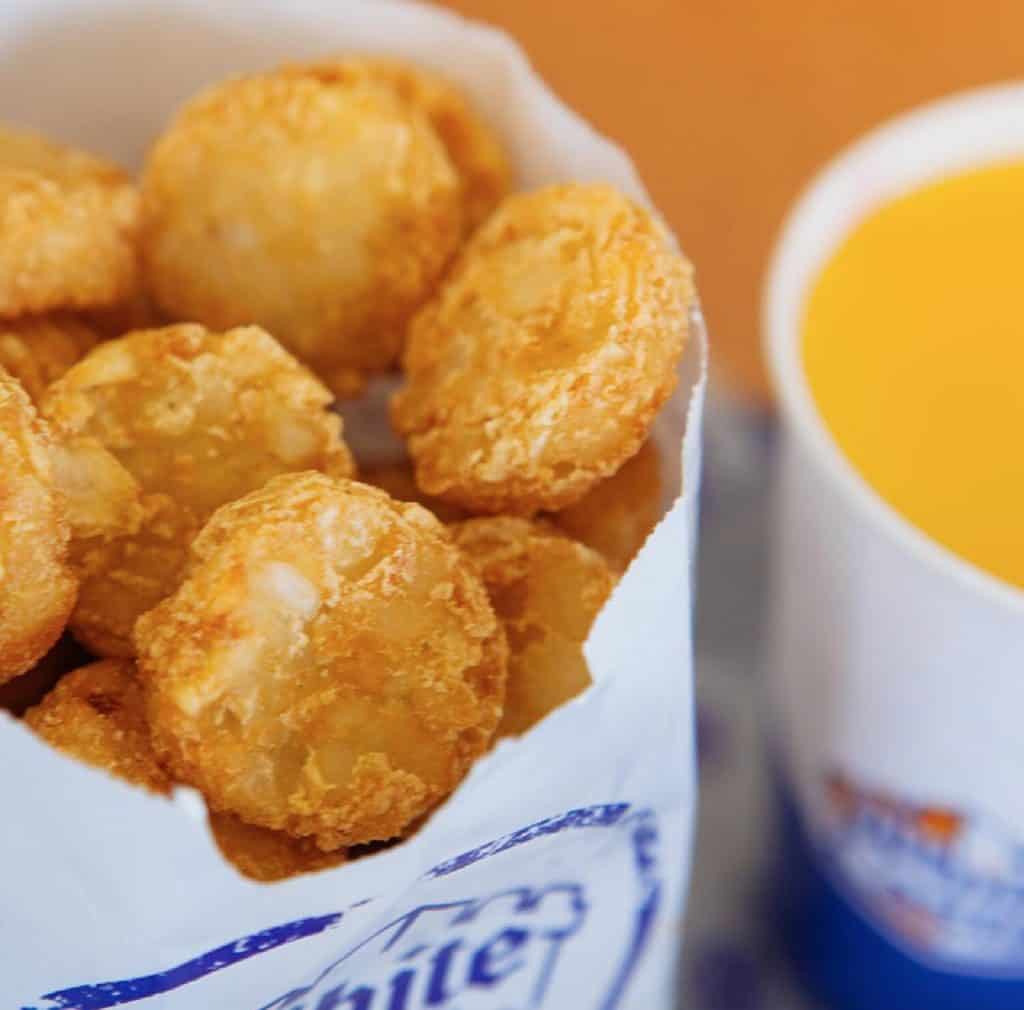 A close up of hash brown rounds in a white and blue White Castle bag alongside a white cup of orange juice.