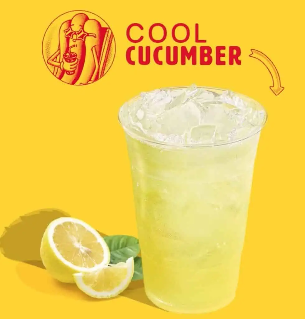 A clear plastic cup filled with cucumber lemonade with slices of lemon and mint sitting near the bottom of the cup with a yellow background to the photo.