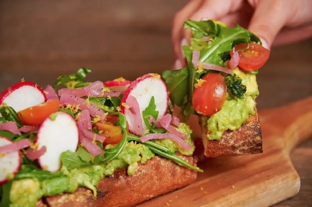 A wooden cutting board with a large, cut loaf of sourdough bread filled with avocado, tomato, greens, carrot, and radish slices.