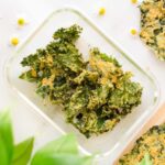 A clear glass container filled with dehydrator kale chips with nutritional yeast.