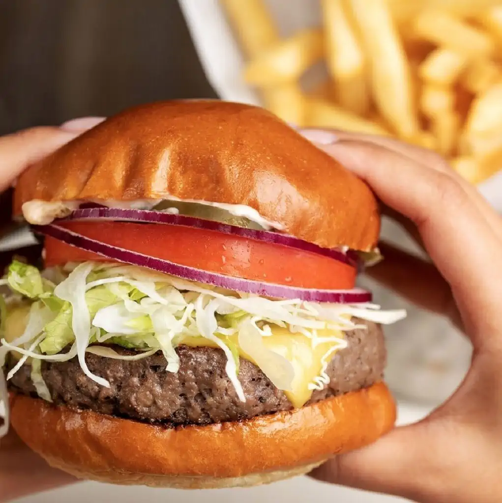 A set of hands holding an Impossible Burger inside of a bun filled with lettuce, vegan cheese, tomato, and onion. Fries are shown in the background.