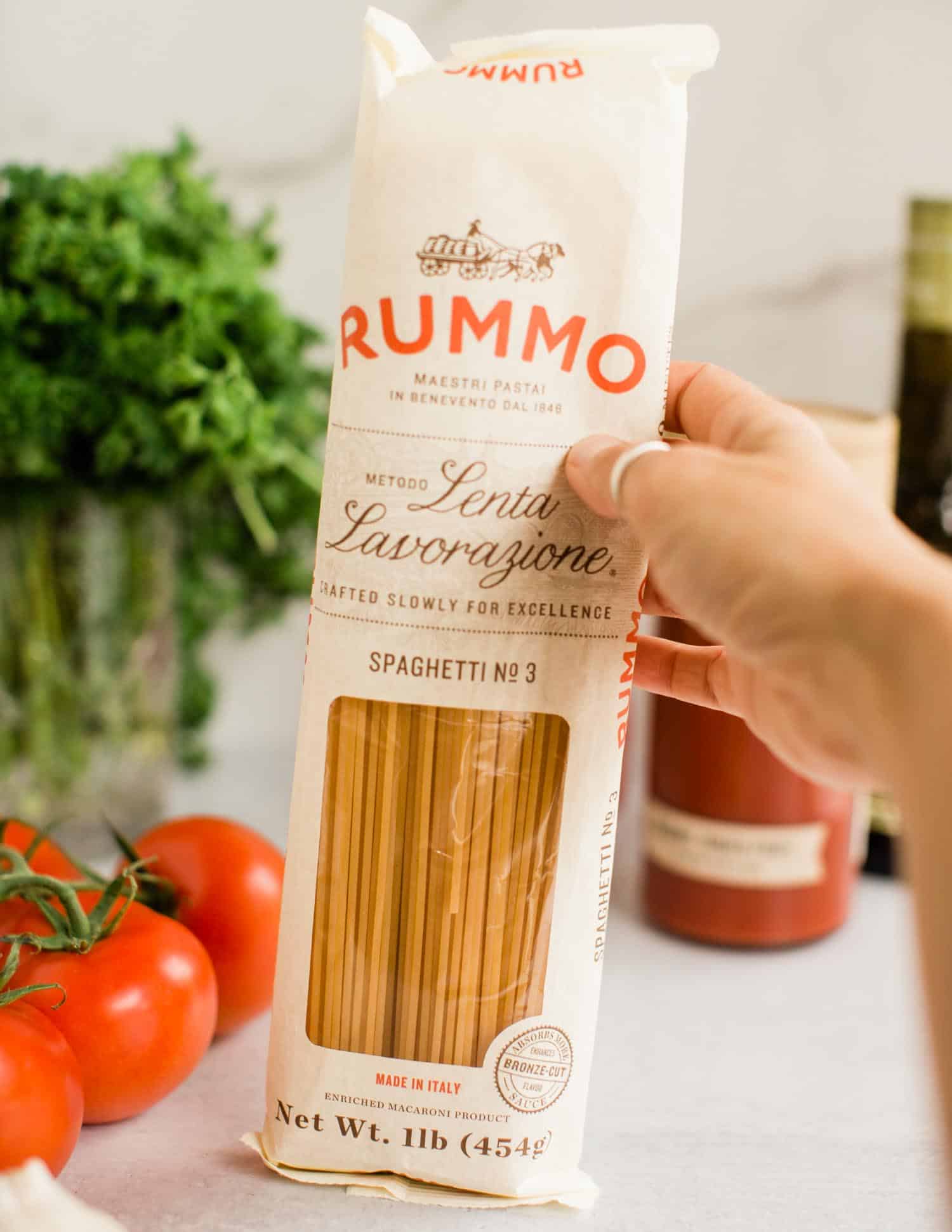 Holding up a package of spaghetti surrounded by tomatoes, parsley, and other traditional Italian ingredients. 